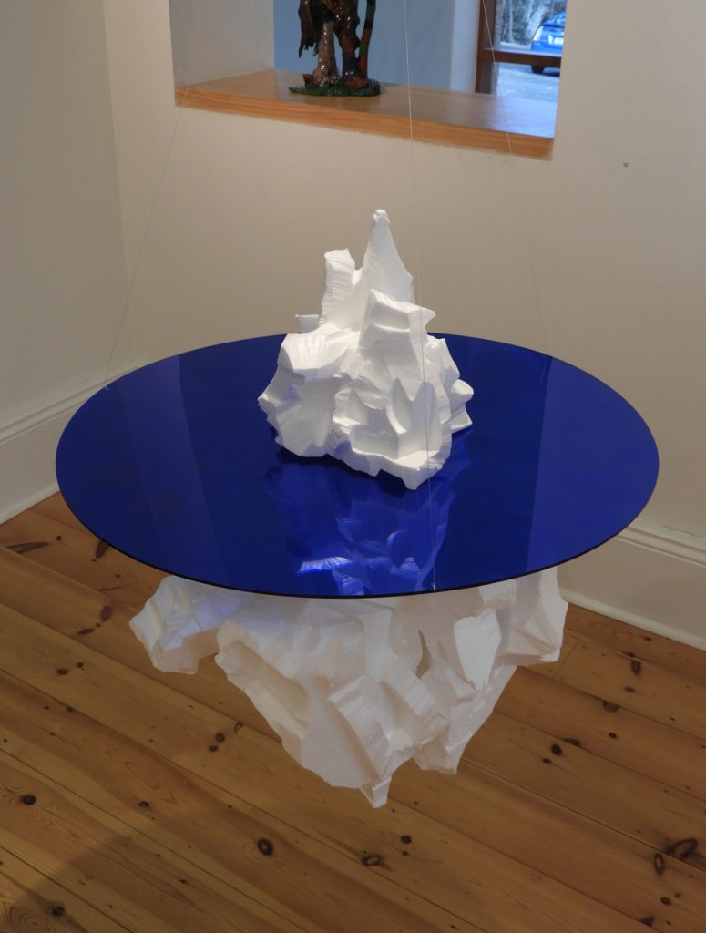 A sculpture of an iceberg cut from styrofoam and mounted through a reflective blue disc of perspex hangs in mid-air in an art gallery